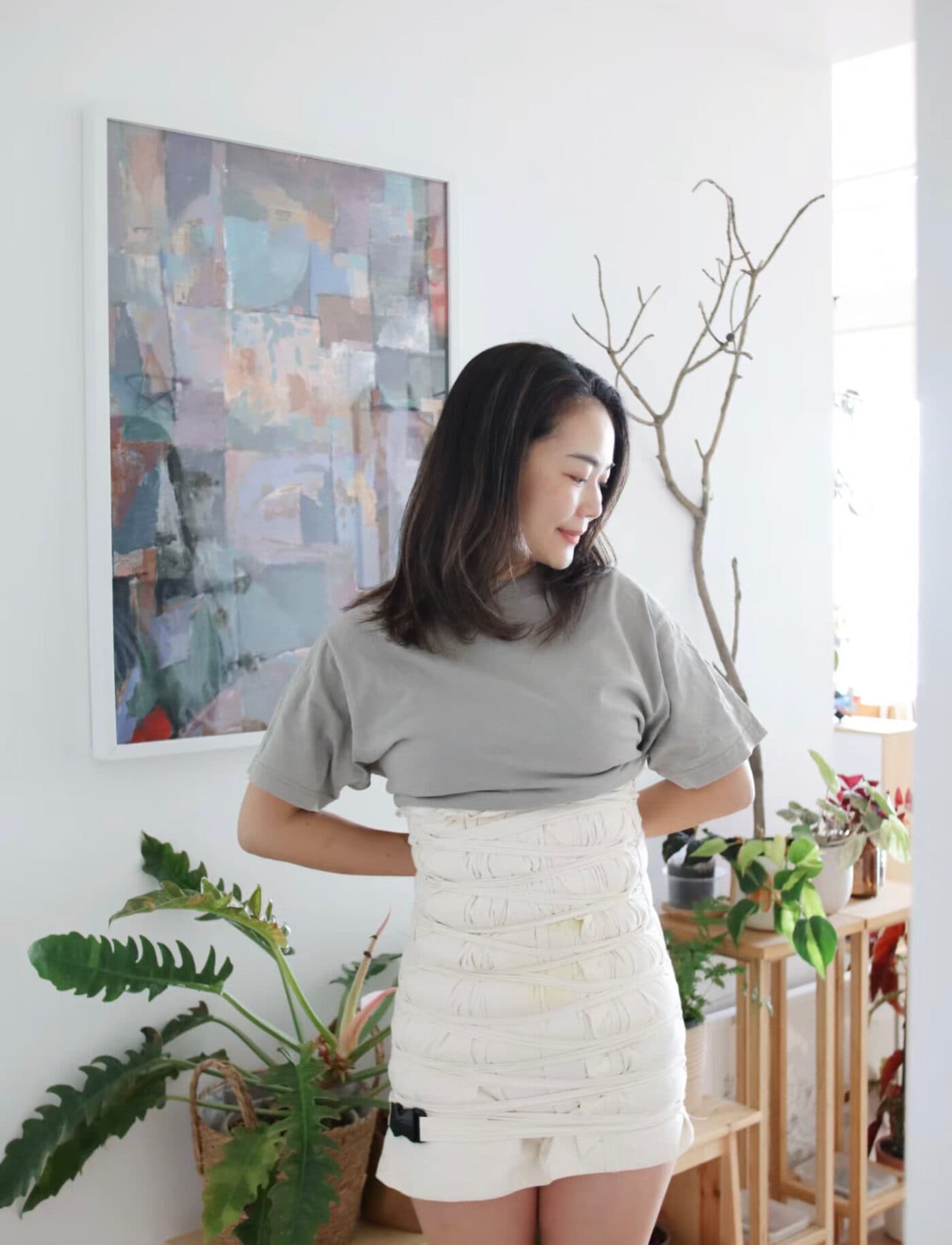 一位妈妈站在自家客厅里,灰色 T 恤下是一圈圈裹好的白色棉布扎肚带,身旁是绿植与木架,墙上挂着一幅抽象画