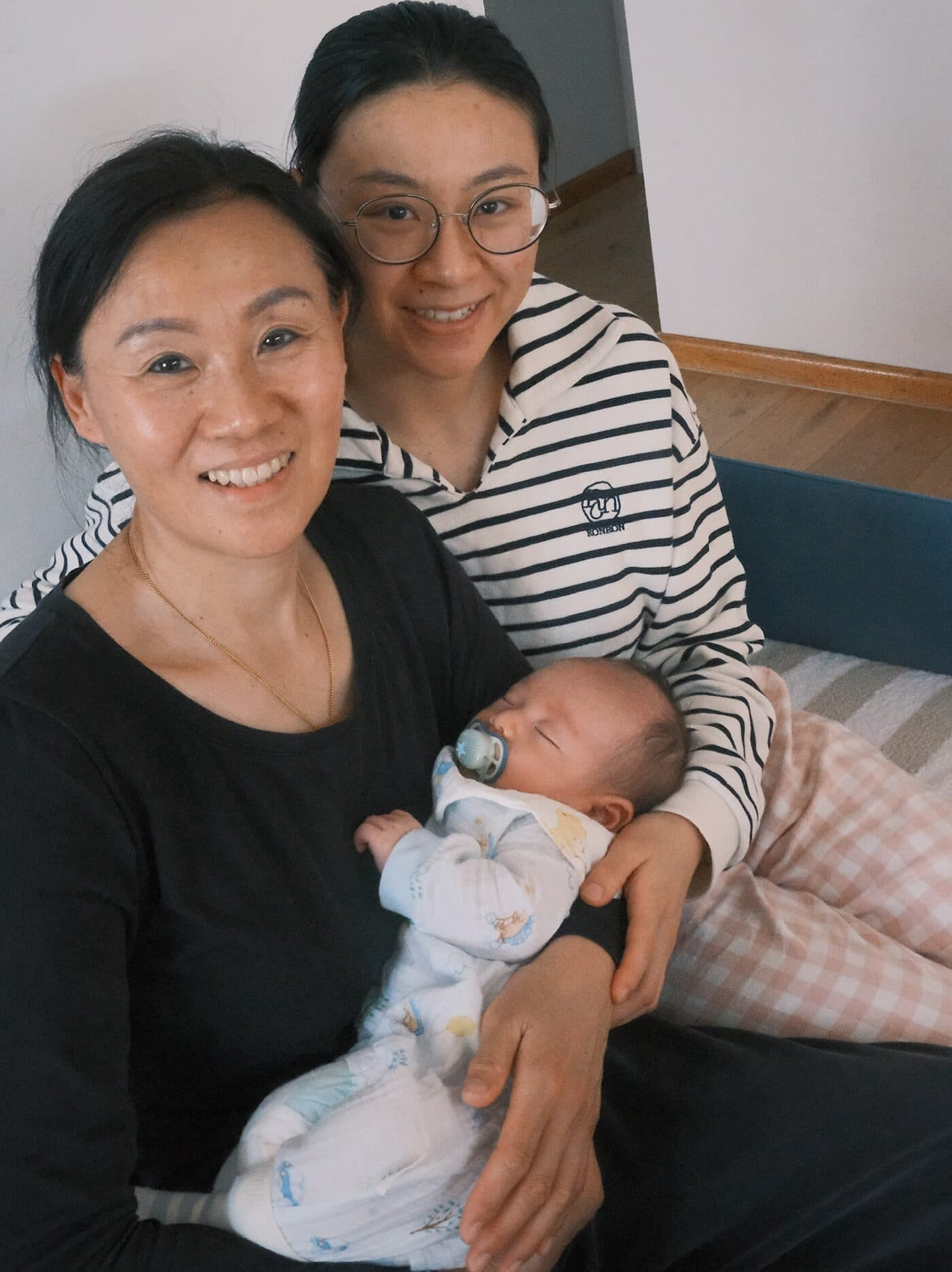 两位女性并肩坐在家中沙发上,怀里抱着含着安抚奶嘴熟睡的新生儿